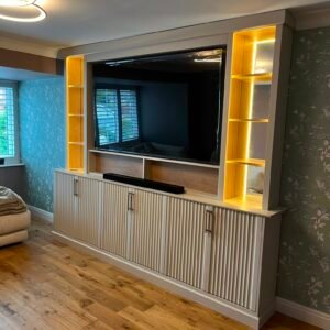 ngled view of a custom living room TV unit showing vertical fluted cabinetry, brass handles, and warm LED strip lighting
