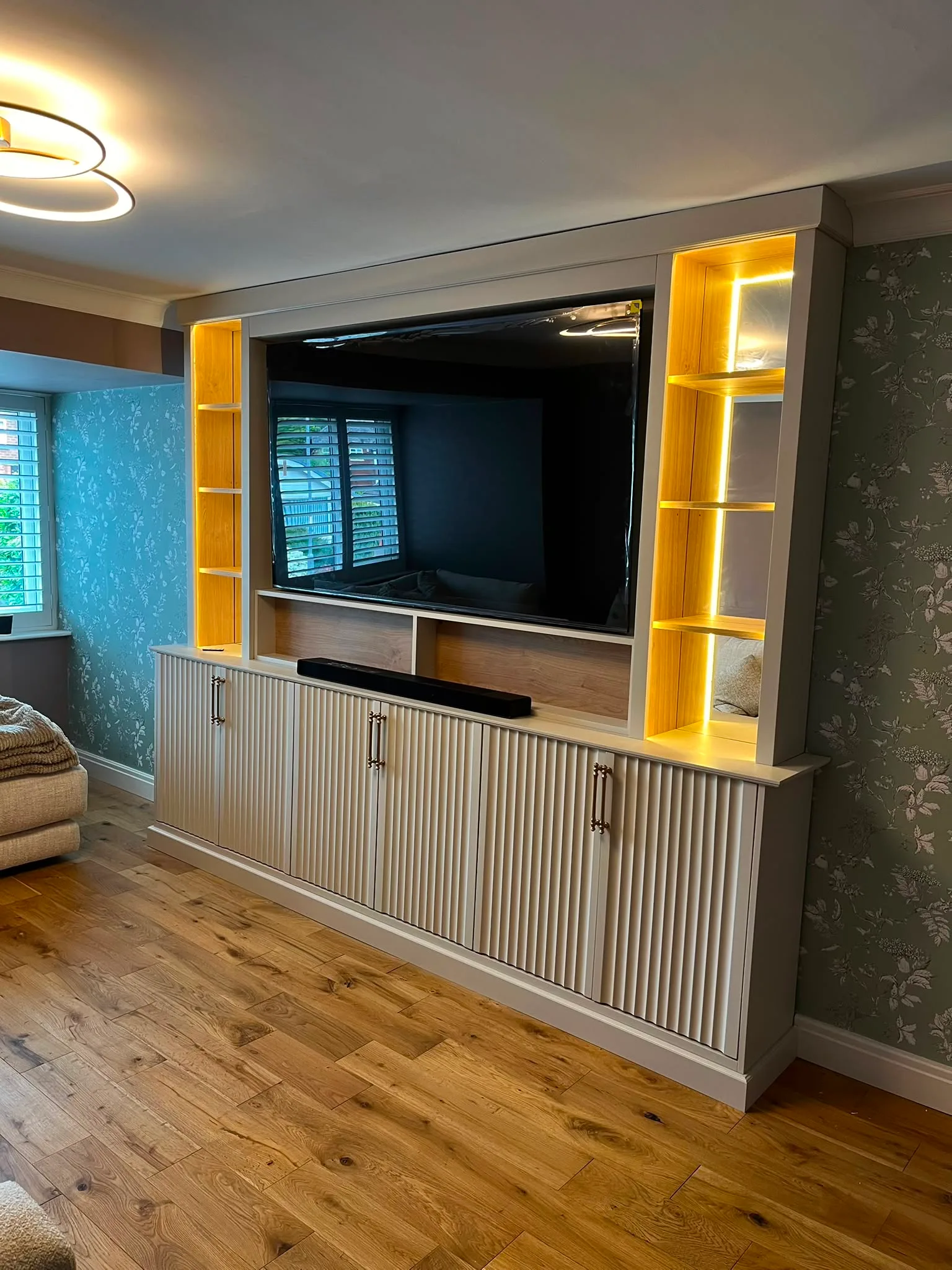 ngled view of a custom living room TV unit showing vertical fluted cabinetry, brass handles, and warm LED strip lighting