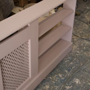 A bespoke, dusty pink painted radiator cover featuring integrated open shelving and traditional lattice fretwork, shown in a busy carpentry workshop.