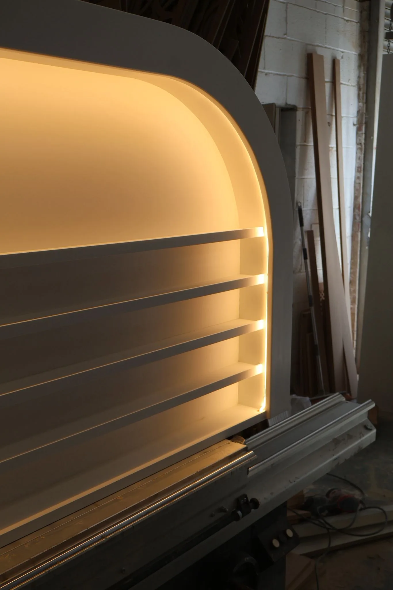 Bespoke white arched wall-mounted display unit with integrated warm LED halo lighting and thin shelves for nail polish bottles in a workshop.