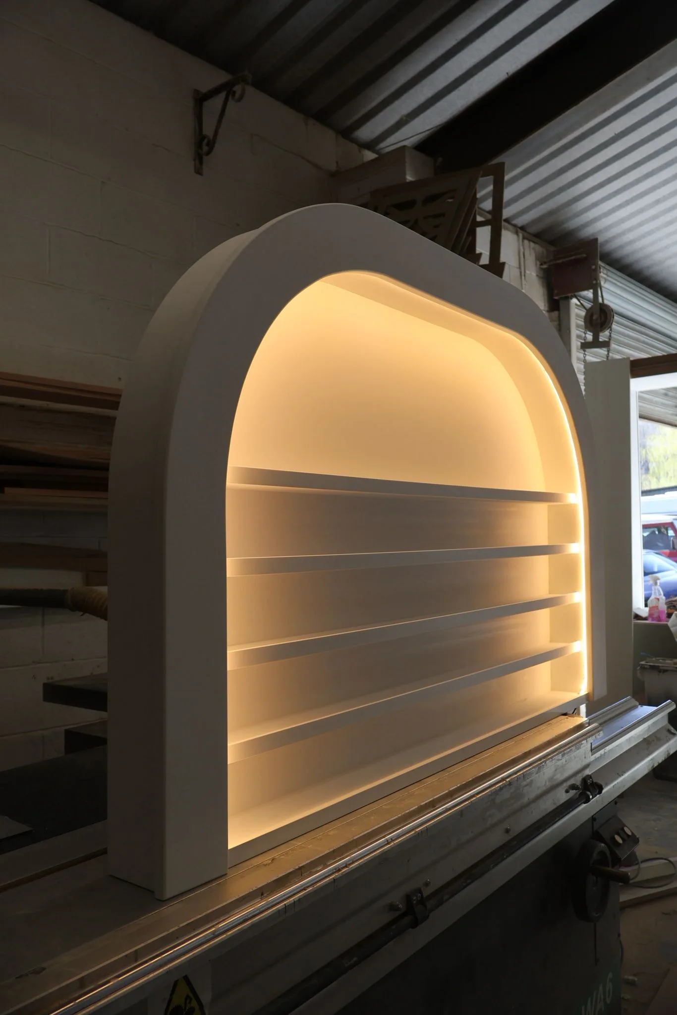 Bespoke white arched wall-mounted display unit with integrated warm LED halo lighting and thin shelves for nail polish bottles in a workshop.